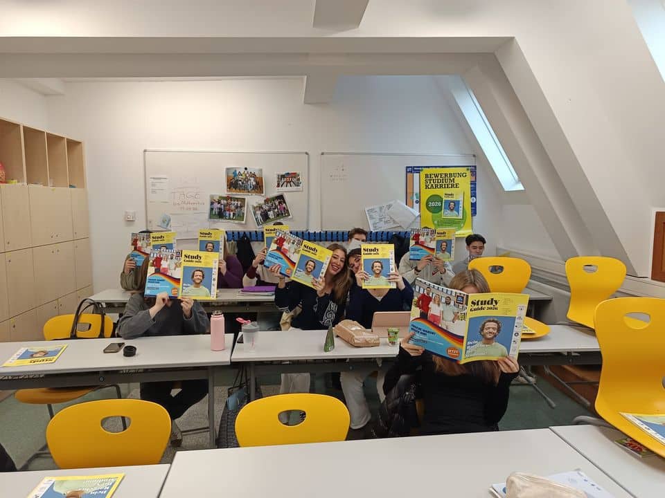 Eine Gruppe von Schülern, die in einem Klassenzimmer an den Tischen sitzen, hält sich Zeitschriften vor das Gesicht, um ihre Identität zu verschleiern. Um den Raum herum sind gelbe Stühle zu sehen.
