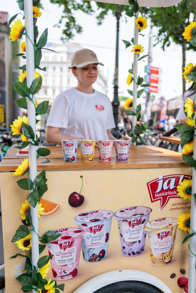 Eine Frau in weißem „Ja! Natürlich“-T-Shirt und Cap steht hinter einem Stand, an dem verschiedene mit Sonnenblumen verzierte Joghurtbecher ausgestellt sind.