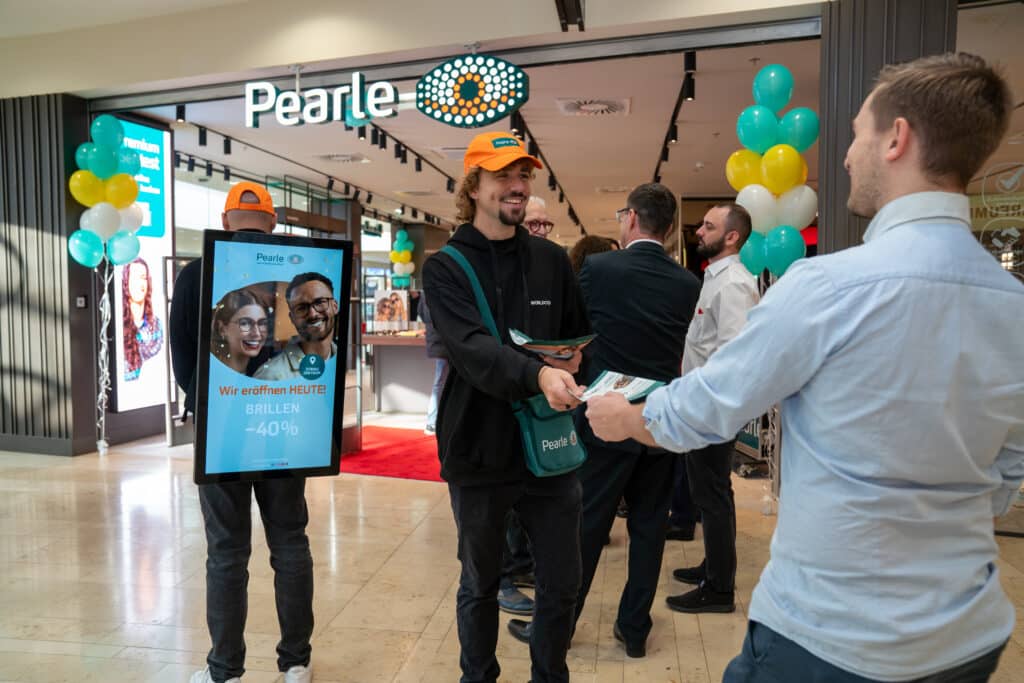 Mehrere Personen stehen vor einem Pearle-Geschäft. Eine Person trägt ein digitales Werbeschild, eine andere reicht einem Mann Flyer. Im Hintergrund sind Luftballons und ein Pearle-Schild zu sehen.