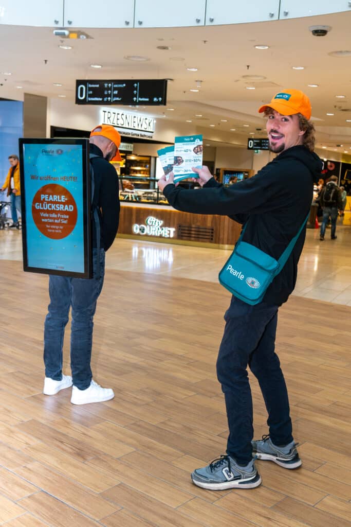 Zwei Promoter mit orangefarbenen Hüten stehen in einem Einkaufszentrum; einer hält Flyer und eine Markentasche, während der andere ein digitales Werbeschild auf dem Rücken trägt.