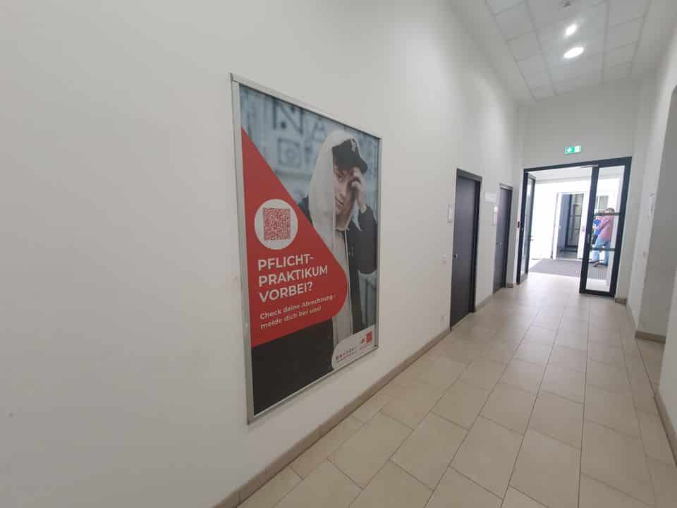 In einem Flur mit gefliestem Boden und weißen Wänden hängt an der linken Wand ein großes deutschsprachiges Praktikumsanzeigenplakat.