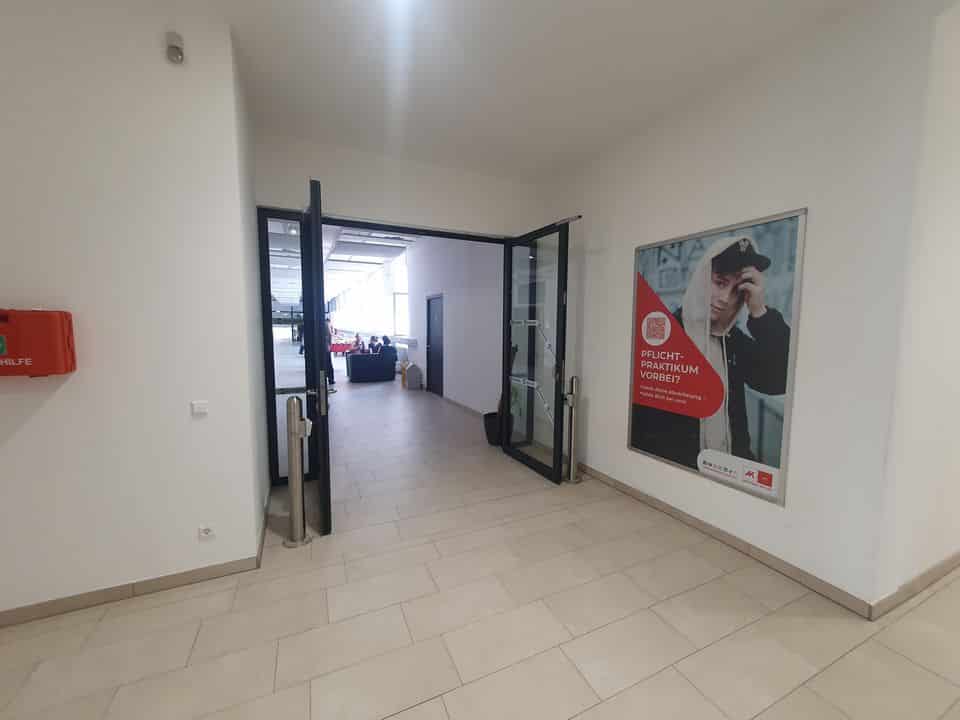 Ein Flur mit offenen Glastüren führt zu einem größeren Raum; an der linken Wand ist ein Feuerlöscher angebracht und an der rechten Wand hängt ein großes Poster.