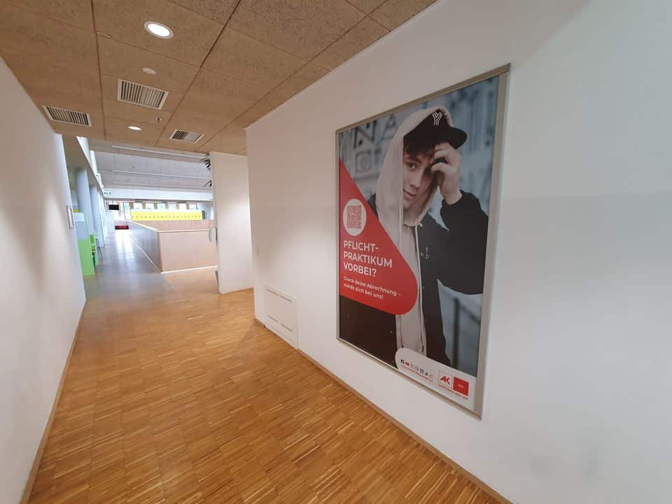 Flur mit Holzboden und weißen Wänden. An der rechten Wand hängt ein großes Poster mit einer Person und Text zum Thema Praktika von der AK Wien