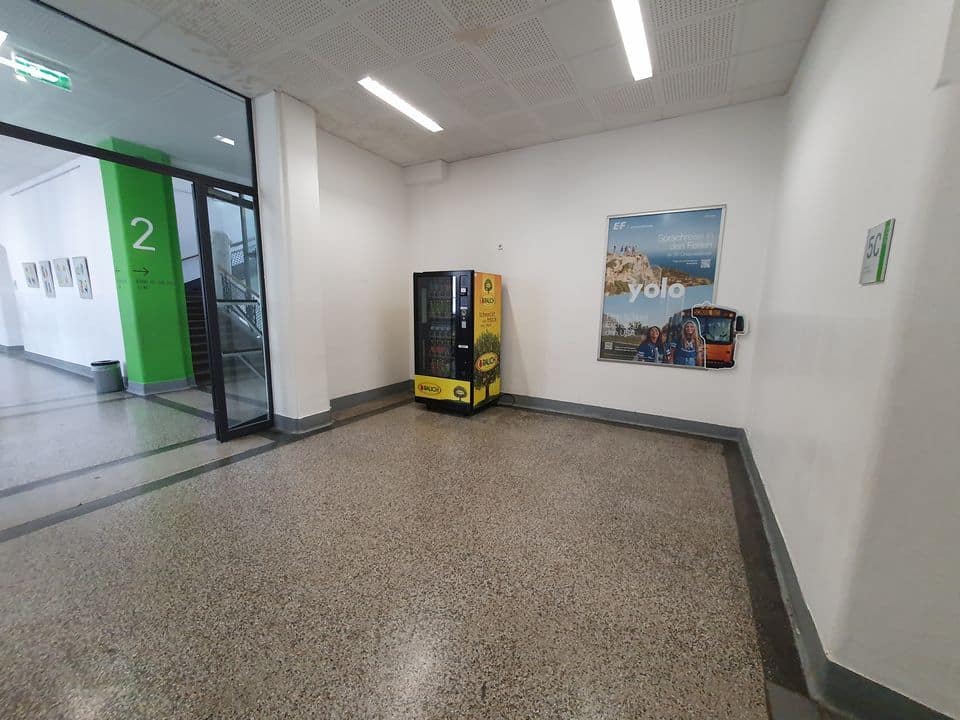 Flur mit einem Getränkeautomaten und einem großen Poster an der Wand. Links befindet sich eine Treppe mit einer Glastür. Der Boden ist braun gesprenkelt, Wände und Decke sind weiß.