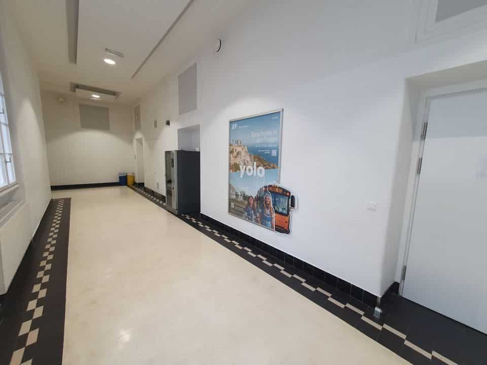 Ein Flur mit weißen Wänden, cremefarbenem Boden und schwarzen Zierleisten. An der Wand hängt ein Poster mit „Yolo“ und einem Busbild. Links ist ein Fenster, rechts eine geschlossene Tür.
