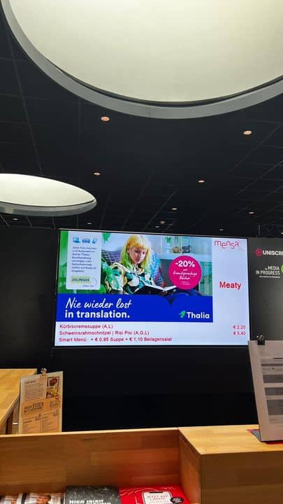 Auf einer digitalen Anzeige hinter einer Theke ist eine Werbung mit einer blonden Frau, die ein Buch liest, und deutschem Text zu sehen; unter der Werbung sind die Preise der Artikel aufgeführt.