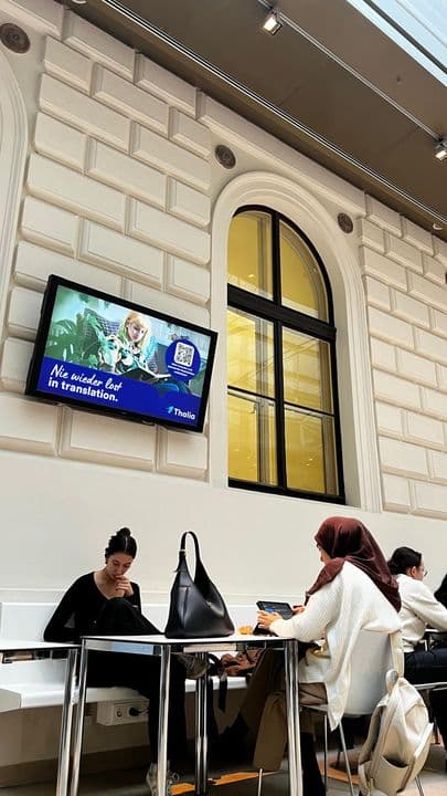 Menschen sitzen und arbeiten an Tischen in einem modernen Gebäude mit einem großen Bogenfenster und einem an der Wand montierten Uniscreen, auf dem eine Werbung angezeigt wird.