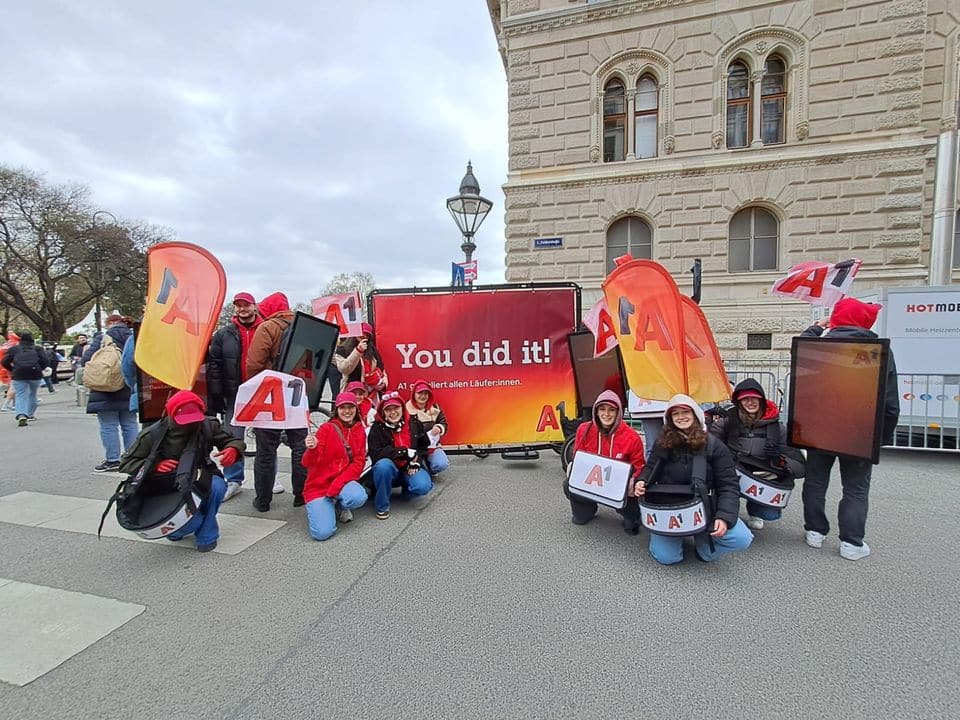 Eine Gruppe von Menschen in passenden rot-blauen Outfits hält „A1“-Schilder und -Flaggen und posiert neben einem Banner mit der Aufschrift „Du hast es geschafft!“ auf einer Stadtstraße.