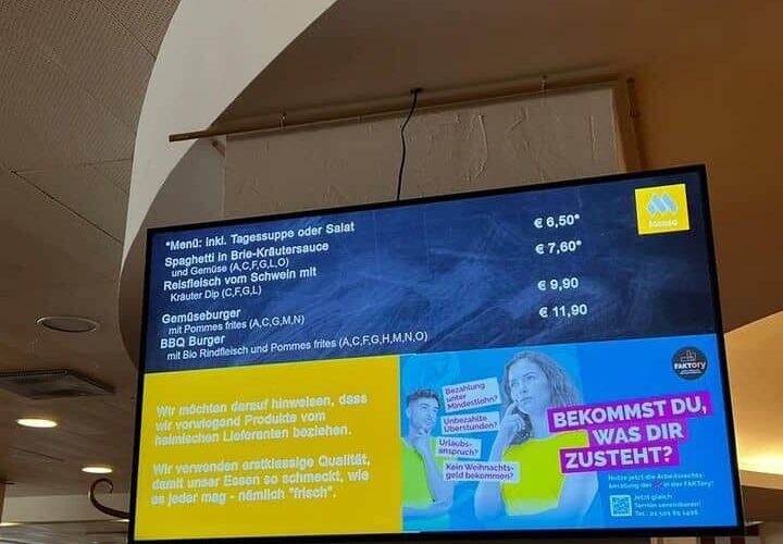 Digitale Speisekarte in einem Restaurant mit verschiedenen Gerichten und Preisen. Text in deutscher Sprache mit gelben und blauen Akzenten. Decke und Leuchten über dem Bildschirm sichtbar.