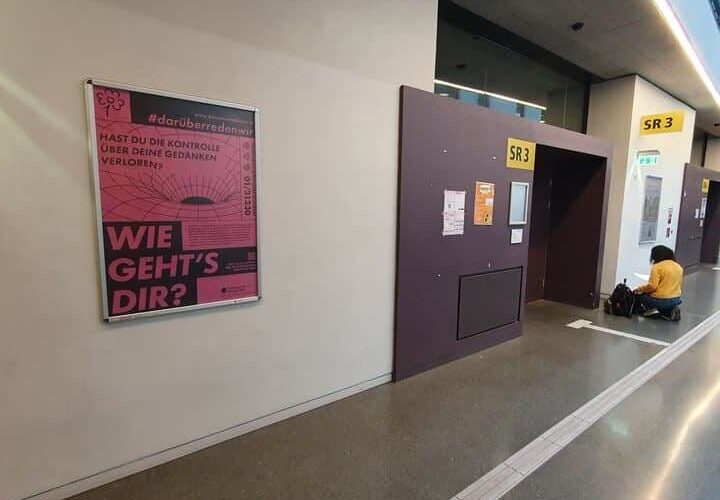 Flur mit einer Person, die auf dem Boden neben Türen mit der Aufschrift SR3 sitzt. Ein rosa Poster an der Wand zeigt deutsche Texte, darunter „Wie geht's dir?“ und „#darüberredenwir“.