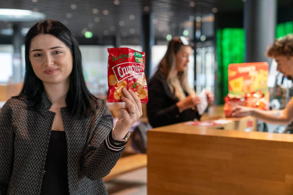 Eine Frau hält bei einer Promotion fürs Marketing in einem modernen Innenraum eine rote Tüte Crunchips-Kartoffelchips hoch, im Hintergrund stehen zwei weitere Personen an einer Theke.