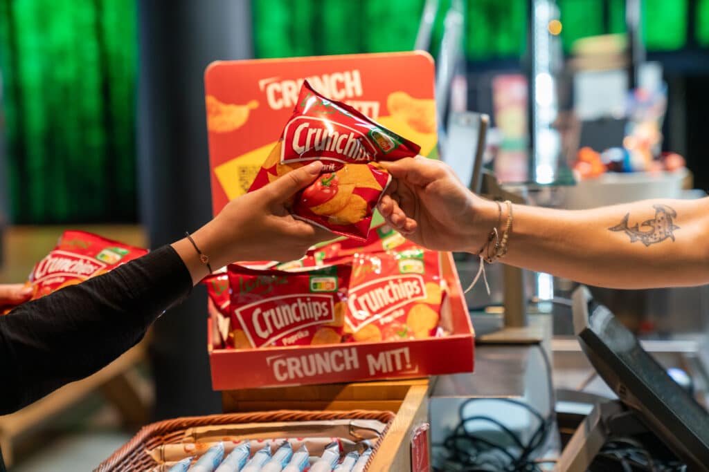 Zwei Personen tauschen eine kleine Tüte Crunchips-Kartoffelchips vor einem mit weiteren Tüten gefüllten Verkaufsständer aus.