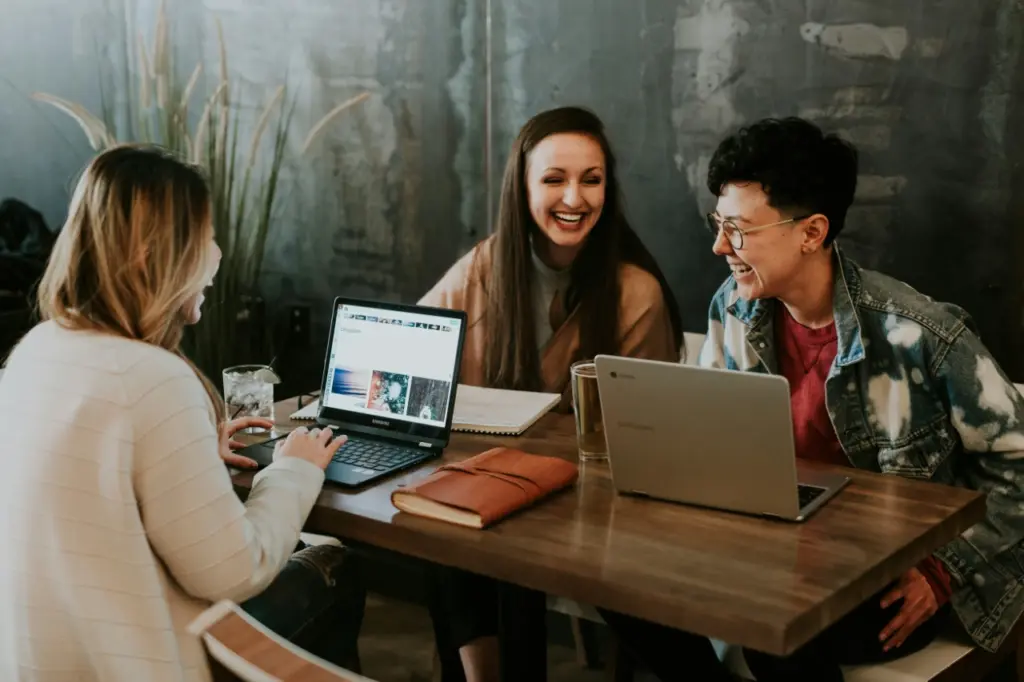 Drei Personen sitzen mit Laptops, Notizbüchern und Getränken an einem Holztisch, unterhalten sich und lächeln – ein ungezwungener Moment, der echte Arbeitszufriedenheit in einer entspannten Innenumgebung widerspiegelt.