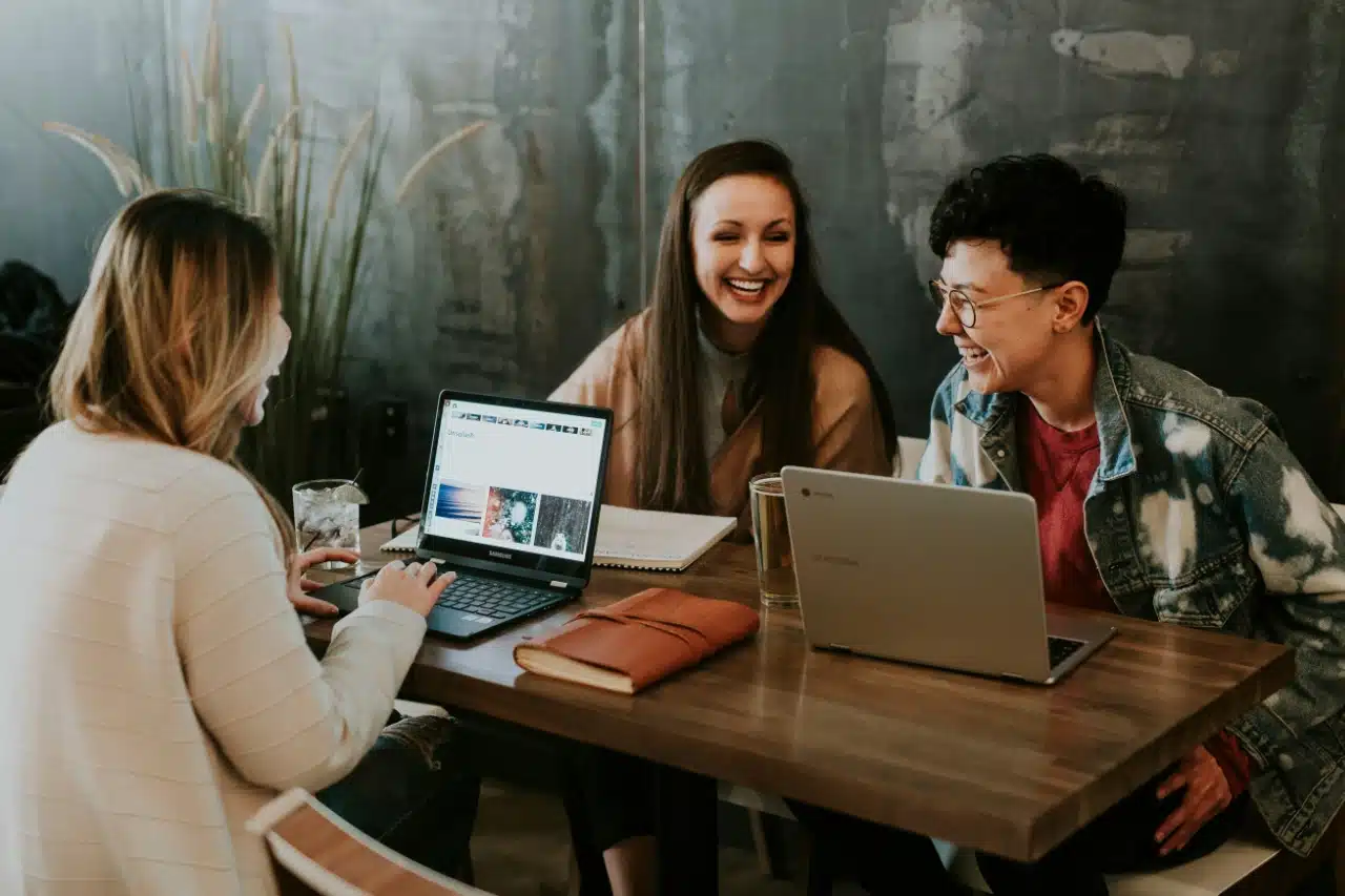 Drei Personen sitzen mit Laptops, Notizbüchern und Getränken an einem Holztisch, unterhalten sich und lächeln – ein ungezwungener Moment, der echte Arbeitszufriedenheit in einer entspannten Innenumgebung widerspiegelt.