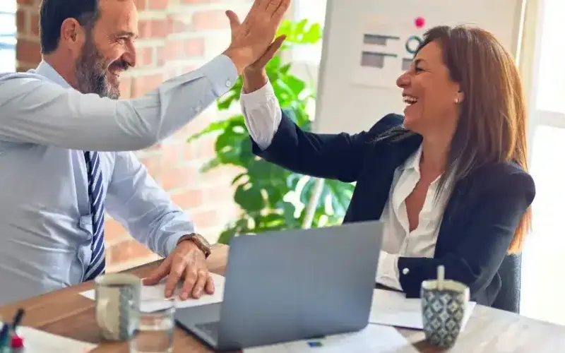 Zwei Geschäftsleute lächeln und geben sich ein High Five an einem Schreibtisch mit einem Laptop, Dokumenten und Kaffeetassen in einer Büroumgebung.