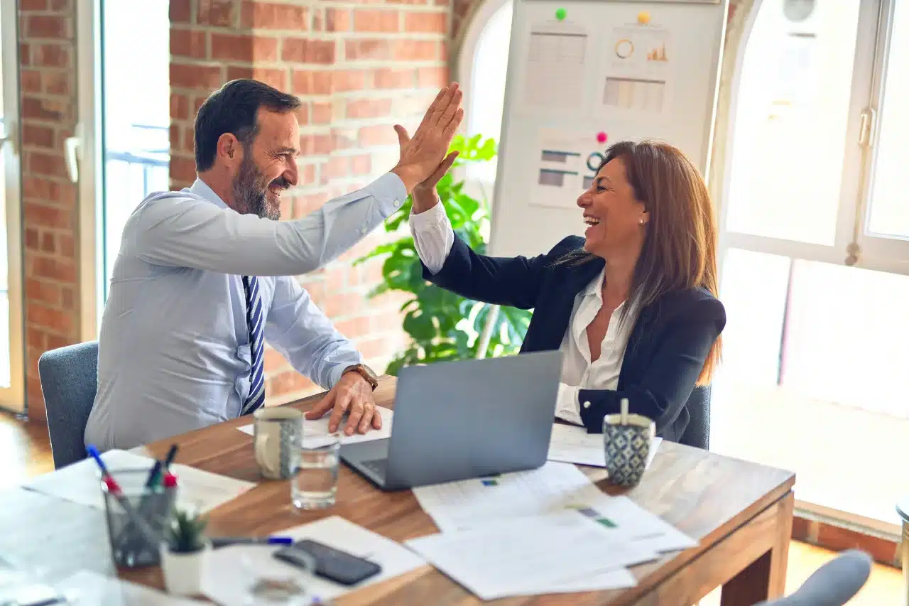 Zwei Geschäftsleute sitzen lächelnd an einem Schreibtisch und geben sich ein High Five. In einer modernen Büroumgebung sind ein Laptop, Dokumente und Diagramme zu sehen – eine Hommage an den Aufbau eines starken Arbeitgeberversprechens.
