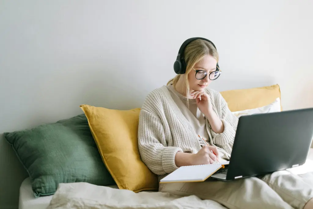 Eine Person mit Kopfhörern aus der Generation Z sitzt auf einem Bett mit Kissen, benutzt einen Laptop und schreibt in ein Notizbuch.