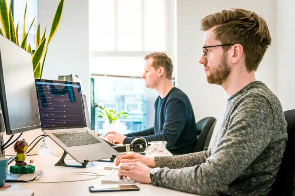Zwei Personen sitzen an Schreibtischen und arbeiten an Computern in einem modernen Büro mit natürlichem Licht und Pflanzen im Hintergrund. Dies schafft eine positive Atmosphäre, die das HR Retention Management unterstützt.
