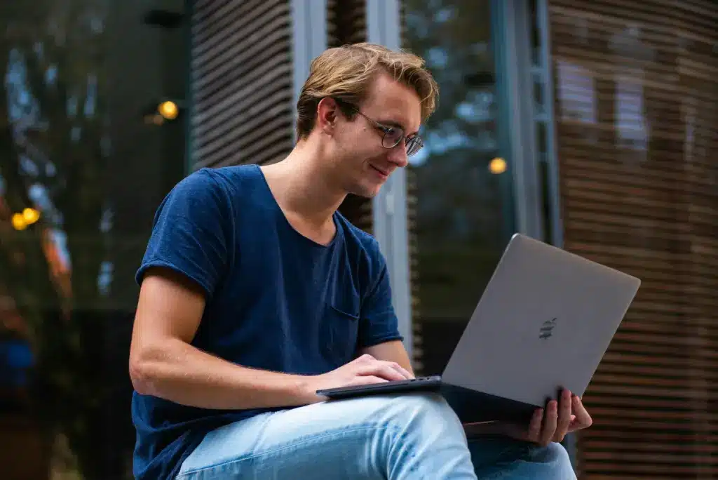 Ein junger Mann der Generation Y bzw. Millennial mit Brille und blauem T-Shirt sitzt lächelnd im Freien und benutzt einen Laptop.
