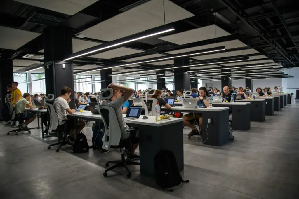 Großes, modernes Büro mit offenen Schreibtischen, an denen die Mitarbeiter an Computern und Laptops arbeiten, was die Zusammenarbeit und effiziente Personalbedarfsplanung am Arbeitsplatz fördert.