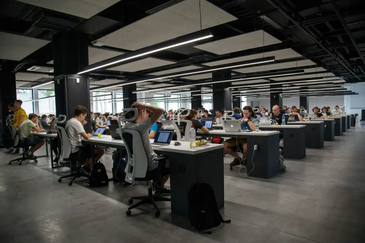 Großes, modernes Büro mit offenen Schreibtischen, an denen die Mitarbeiter an Computern und Laptops arbeiten, was die Zusammenarbeit und effiziente Personalbedarfsplanung am Arbeitsplatz fördert.