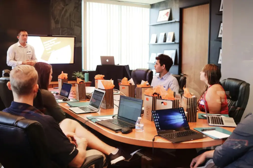 Eine Gruppe von Menschen sitzt mit Laptops und Geschenktüten um einen Konferenztisch herum und hört einem Vortragenden zu, der neben einer Leinwand, auf der eine Präsentation läuft, über Personalentwicklung spricht.