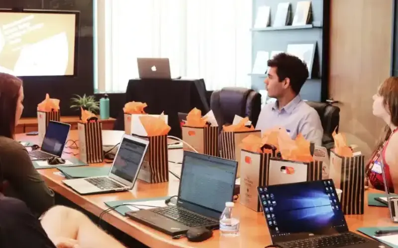 Eine Gruppe von Menschen sitzt mit Laptops und Geschenktüten um einen Konferenztisch und sieht sich in einem modernen Büro eine Präsentation auf einem großen Bildschirm an.