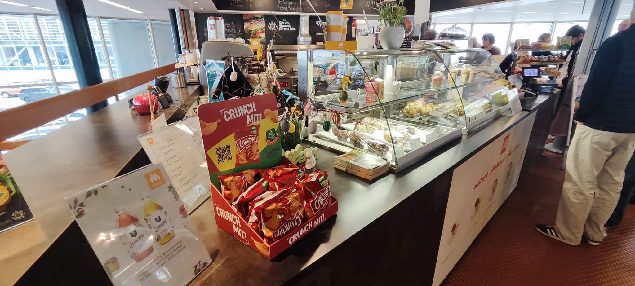 Eine Café-Theke mit einer Dessertvitrine, einem Korb mit Crunchips-Snacks, einem Menüständer und Kunden, die Schlange stehen, um zu bestellen.