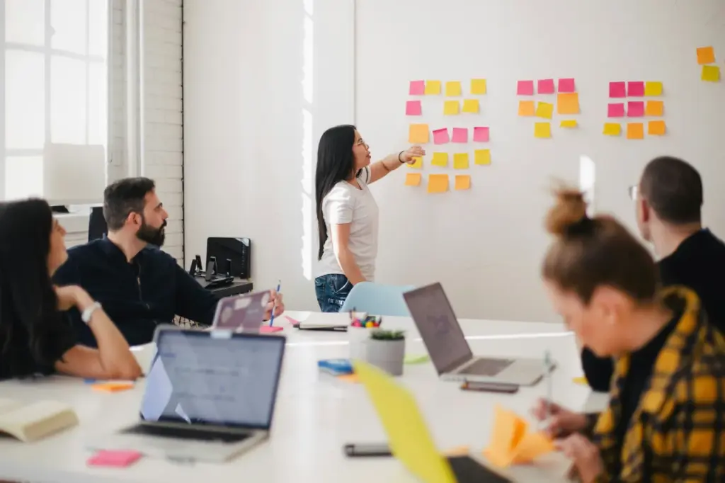 Vier Personen sitzen mit Laptops an einem Tisch, während eine Person auf Haftnotizen auf einem Whiteboard zeigt und so das Alleinstellungsmerkmal der Gruppe in einem hellen Konferenzraum hervorhebt.
