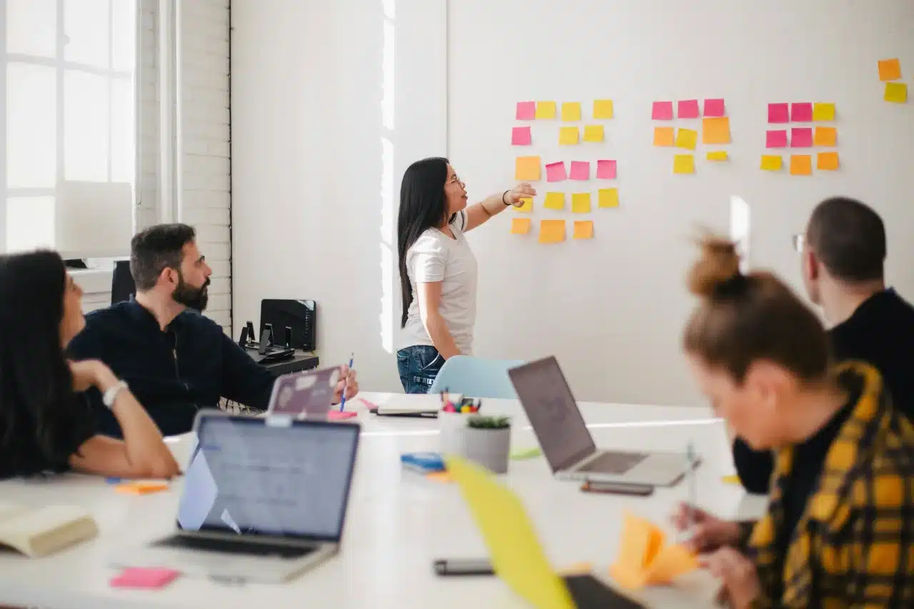 Vier Personen sitzen mit Laptops an einem Tisch, während eine Person auf Haftnotizen auf einem Whiteboard zeigt und so das Alleinstellungsmerkmal der Gruppe in einem hellen Konferenzraum hervorhebt.