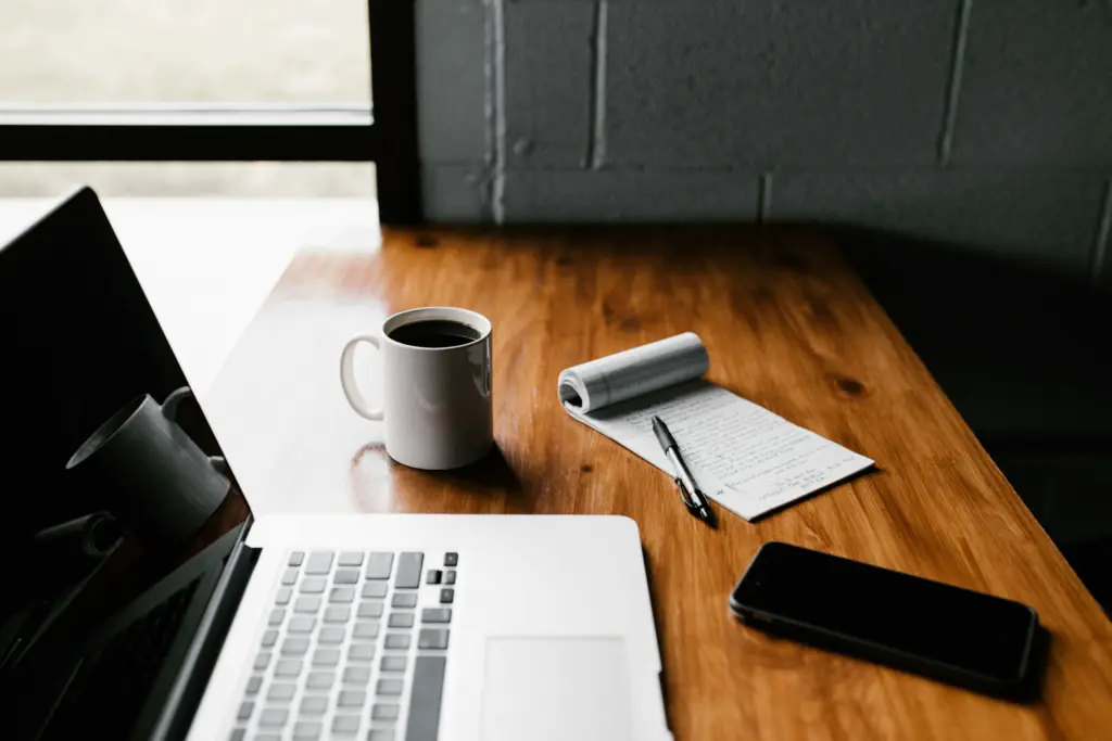 Ein Laptop, eine Tasse Kaffee, ein Notizblock mit handschriftlichen Notizen zu einem Bewerbungsgespräch, ein Stift und ein Smartphone auf einem Holztisch am Fenster.