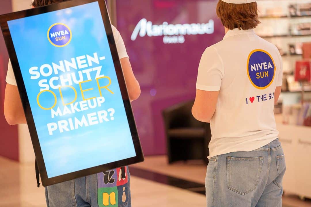 Zwei Personen in Nivea-Sun-Shirts stehen in einem Einkaufszentrum; eine trägt ein digitales Schild mit der Aufschrift "SONNENSCHUTZ ODER MAKEUP PRIMER?" auf Deutsch.