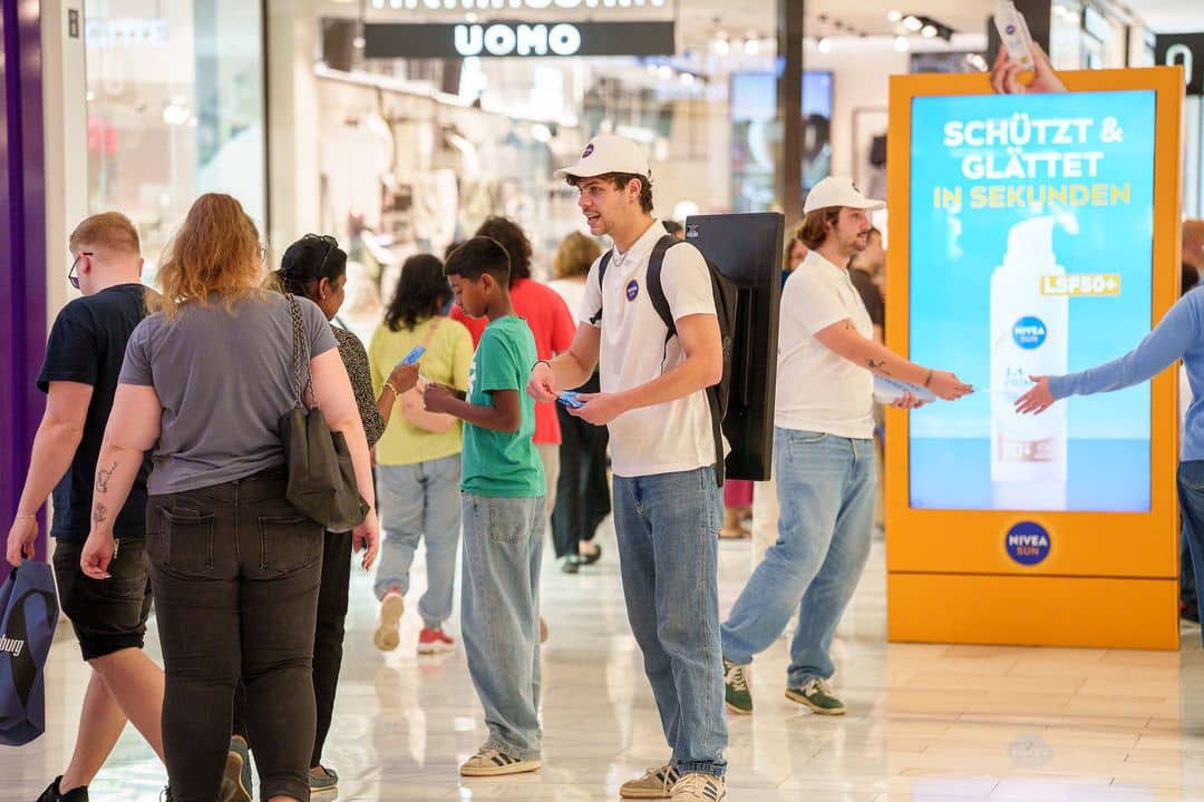 Menschen gehen in einem Einkaufszentrum spazieren, während zwei Promoter mit weißen Hemden und Mützen in der Nähe eines Nivea-Werbedisplays Flugblätter verteilen.