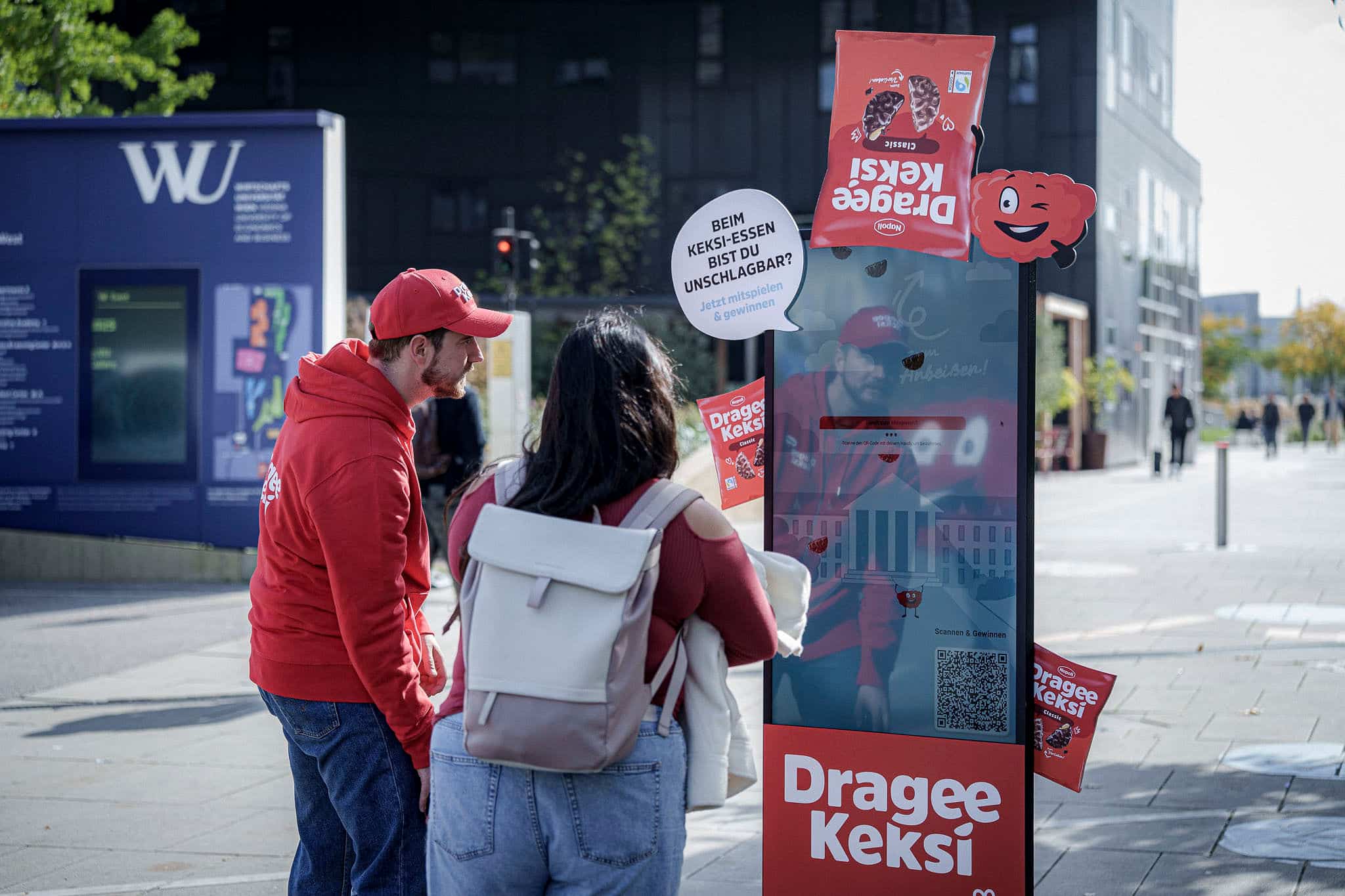 Zwei Personen stehen vor einem Werbeständer für Dragee Keksi auf einem Bürgersteig in der Stadt, mit Plakaten und einem Sprechblasenschild in deutscher Sprache.