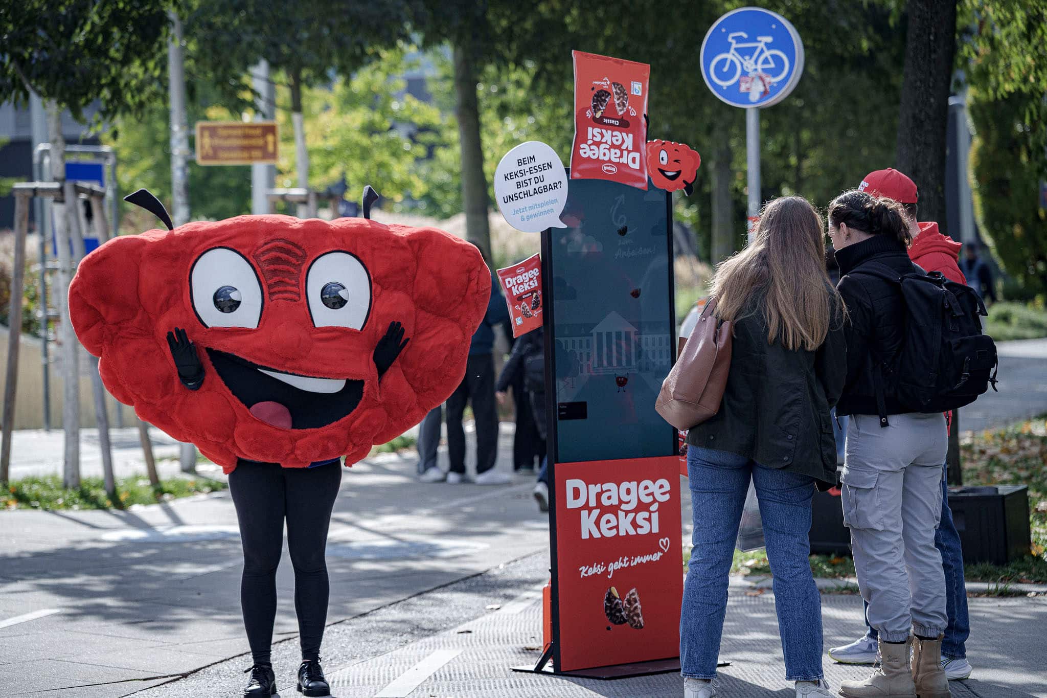 Eine Person in einem roten Maskottchen-Kostüm steht in der Nähe eines Werbekiosks, während eine Gruppe von Menschen auf einem städtischen Bürgersteig mit dem Kiosk interagiert.