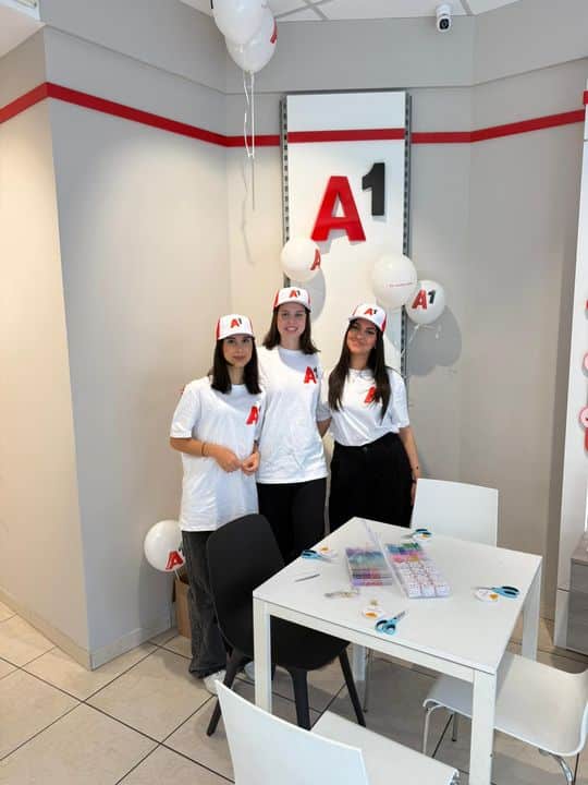 Drei Frauen mit weißen "A1"-Shirts und -Hüten stehen zusammen in einem modernen, weißen Raum, der mit "A1"-Luftballons und Branding dekoriert ist.