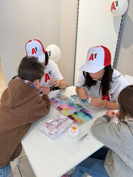 Zwei Erwachsene und zwei Kinder sitzen an einem Tisch und basteln Perlen. Die Erwachsenen tragen A1-Hüte und Hemden, und auf dem Tisch sind bunte Perlen verteilt.