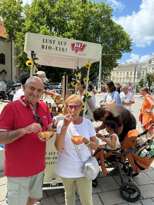 Zwei Menschen essen Eis vor einem "Lust auf Bio-Eis?"-Wagen auf einem Markt im Freien, mit anderen Menschen und einem Kinderwagen im Hintergrund an einem sonnigen Tag.