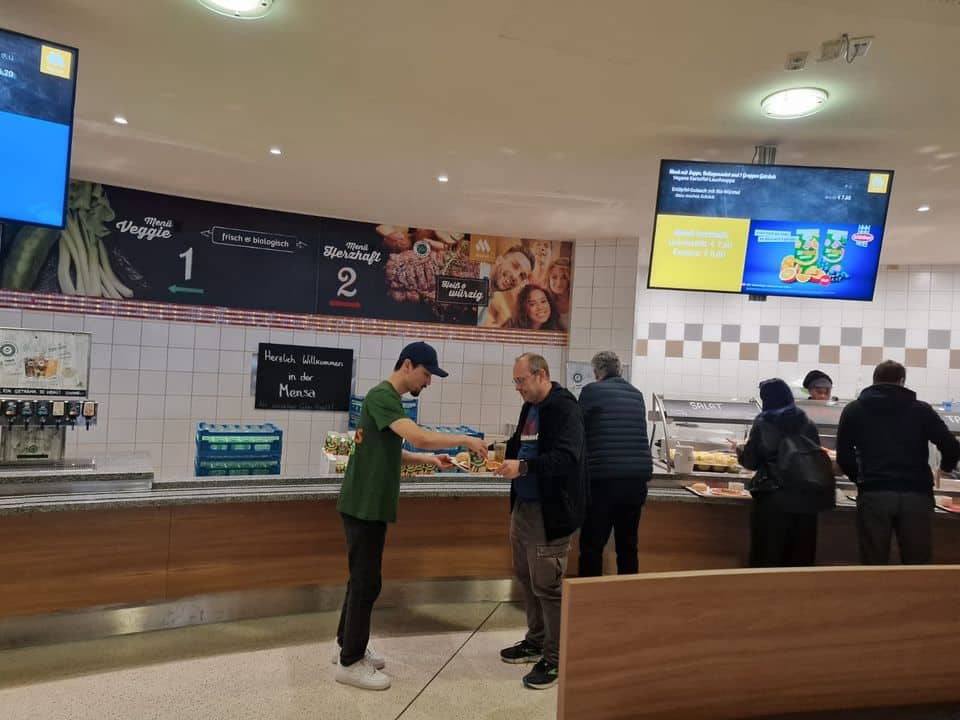 Zwei Männer stehen an einer Cafeteria-Theke, während der eine dem anderen das Essen reicht; im Hintergrund bedienen sich andere Personen am Buffet.