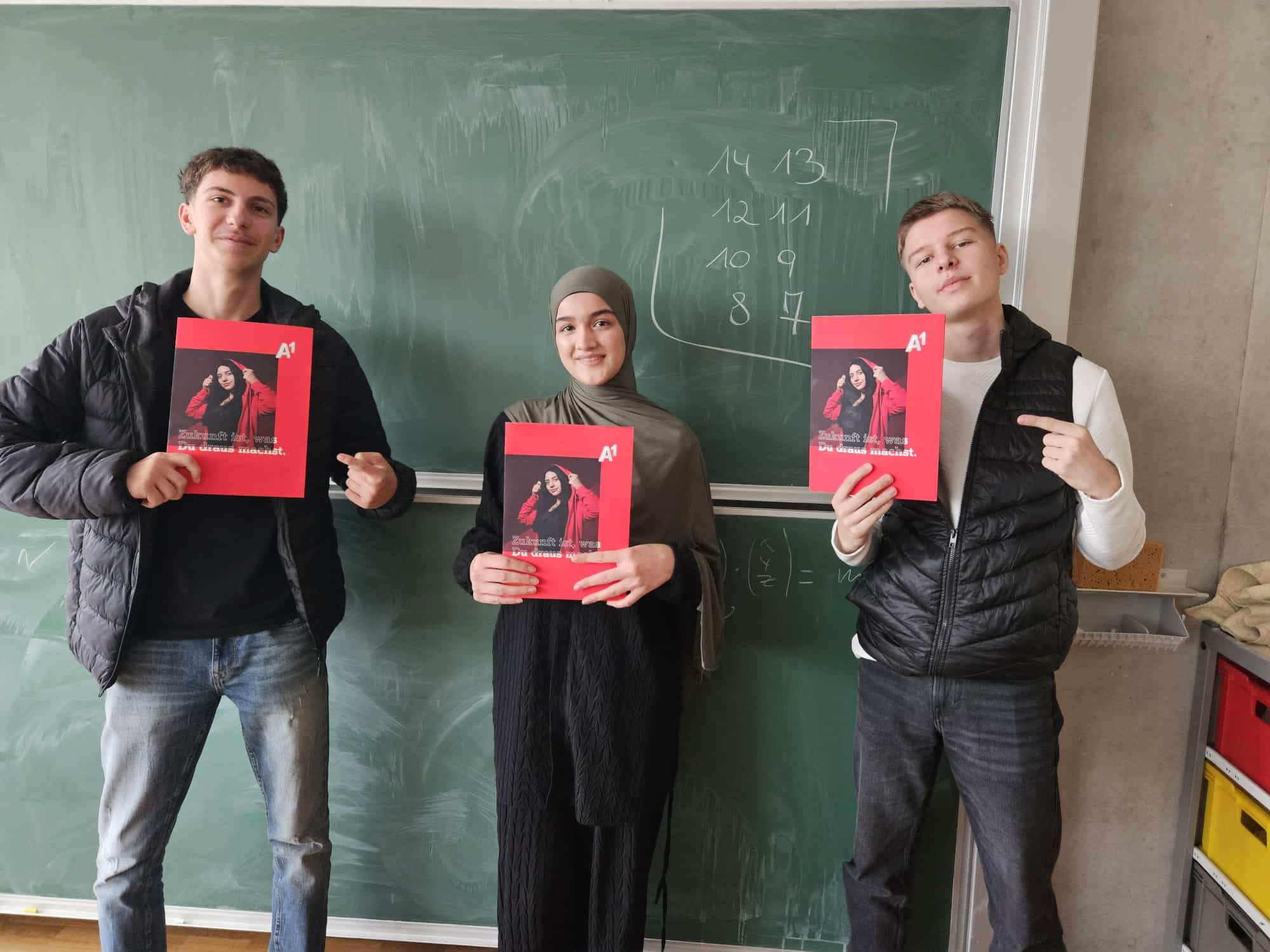 Drei Jugendliche stehen vor einer Kreidetafel, jeder hält eine rote Mappe mit einem Foto in der Hand und schaut in die Kamera.