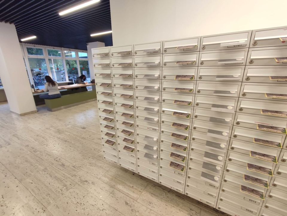 Reihen von weißen Briefkästen säumen einen Flur in einem modernen Gebäude; an den Fenstern im Hintergrund sitzen und arbeiten Menschen an Tischen.