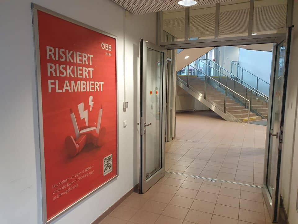 Ein Flur mit einer offenen Glastür, die zu einer Treppe führt; ein rotes Plakat an der Wand warnt auf Deutsch vor den Risiken von E-Scooter-Batterien.