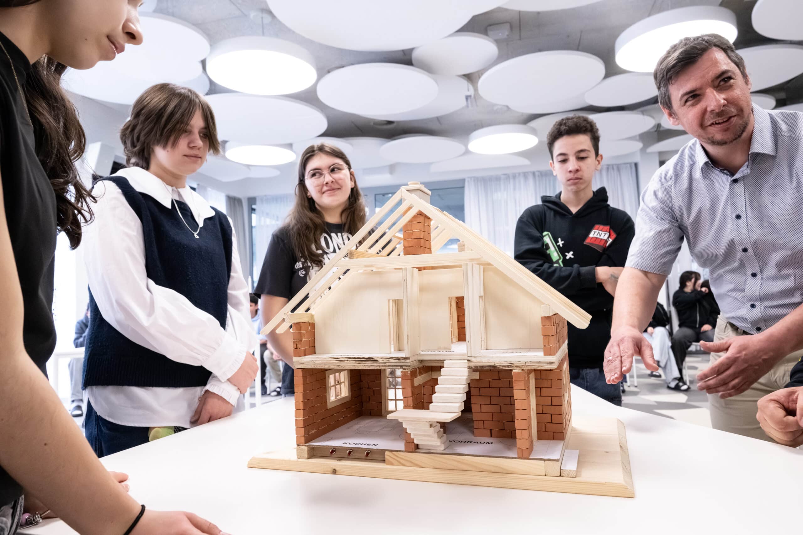 Eine Gruppe von Schülern und ein Lehrer stehen um ein detailliertes Architekturmodell eines Hauses auf einem Tisch in einem modernen Klassenzimmer.