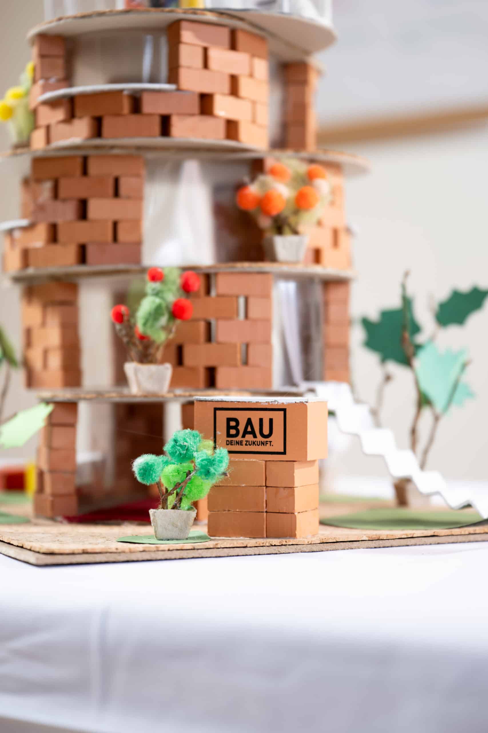 Ein Modell eines runden Backsteingebäudes mit kleinen Pflanzendekorationen und einem Karton mit der Aufschrift "BAU DEINE ZUKUNFT", der im Vordergrund steht.