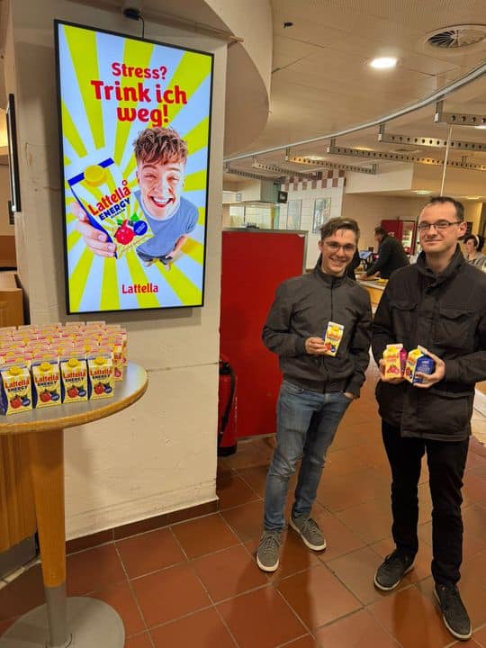 Zwei Personen stehen in einem Raum mit Latella-Getränken neben einer Auslage mit Latella-Flaschen, während hinter ihnen auf einem Bildschirm eine Werbung für Latella zu sehen ist.