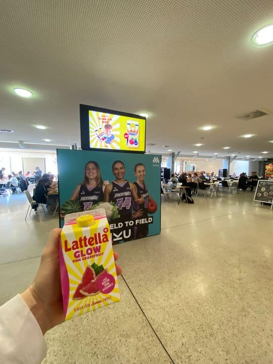 Eine Hand hält eine Packung Lattella Glow vor dem Hintergrund eines Plakats, das drei Sportler in einer belebten Cafeteria zeigt.
