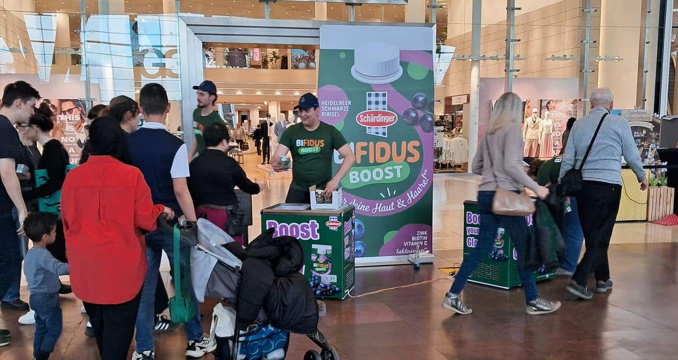 In einem Einkaufszentrum versammeln sich Menschen an einem Werbestand für Schärdingers „Bifidus Boost“, an dem Mitarbeiter Kostproben und Informationsmaterial verteilen.