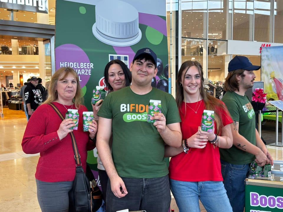 Vier Personen stehen in einem Raum und halten Flaschen mit probiotischen Getränken in der Hand, hinter ihnen ist ein Werbeständer zu sehen. Die Person in der Mitte trägt ein „Bifido Boost“-T-Shirt und eine Kappe.
