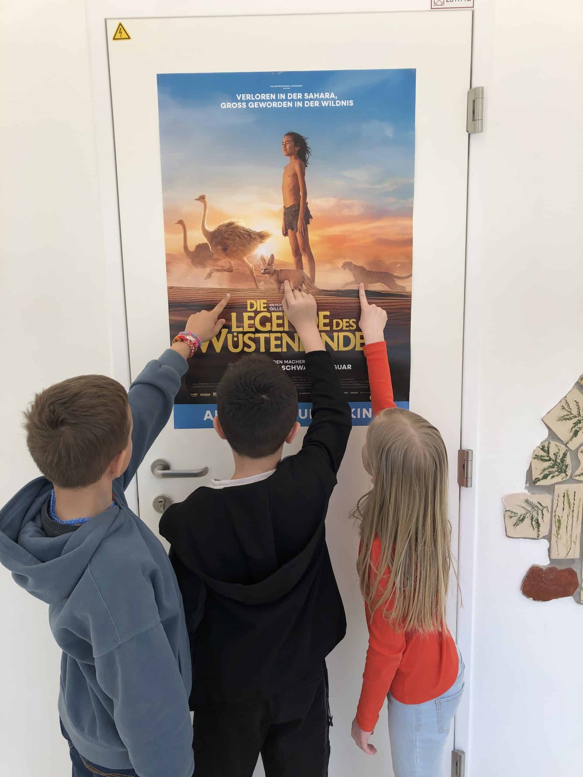 Drei Kinder stehen vor einer weißen Tür und zeigen auf ein buntes Filmplakat, auf dem ein Mädchen und Strauße in einer Wüstenkulisse zu sehen sind.