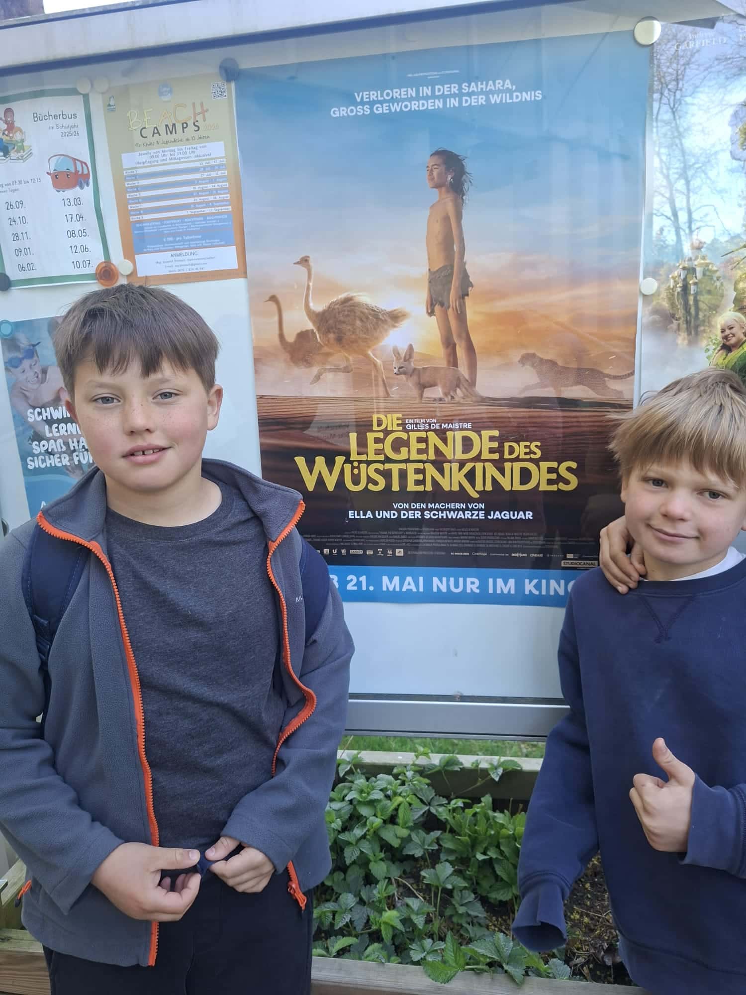 Zwei Jungen stehen vor einer Pinnwand, an der ein deutsches Filmplakat für „Die Legende des Wüstenkindes“ hängt.